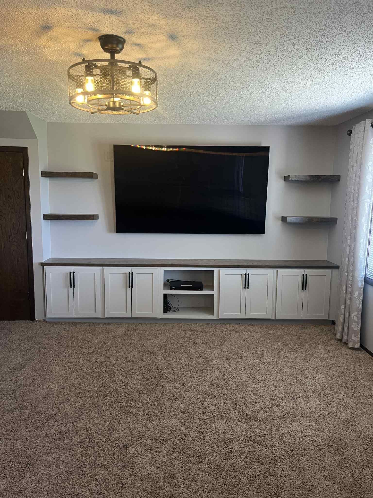 Living room with a large TV above white cabinets, shelves, and a patterned carpet.