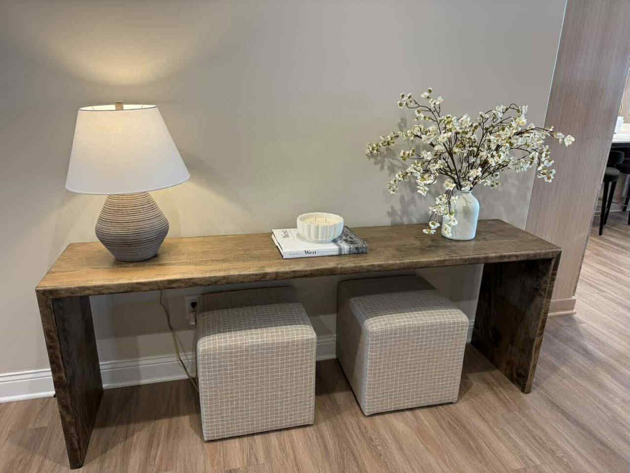 Wooden console table with lamp, flowers, books, and two square ottomans. Light wood floors, neutral walls.