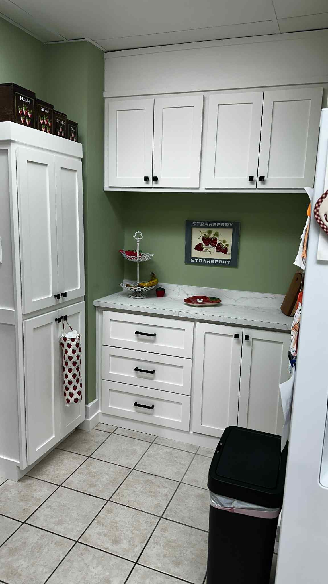 White pantry with cabinets, drawers, and a tall pantry against a green wall and tiled floor.