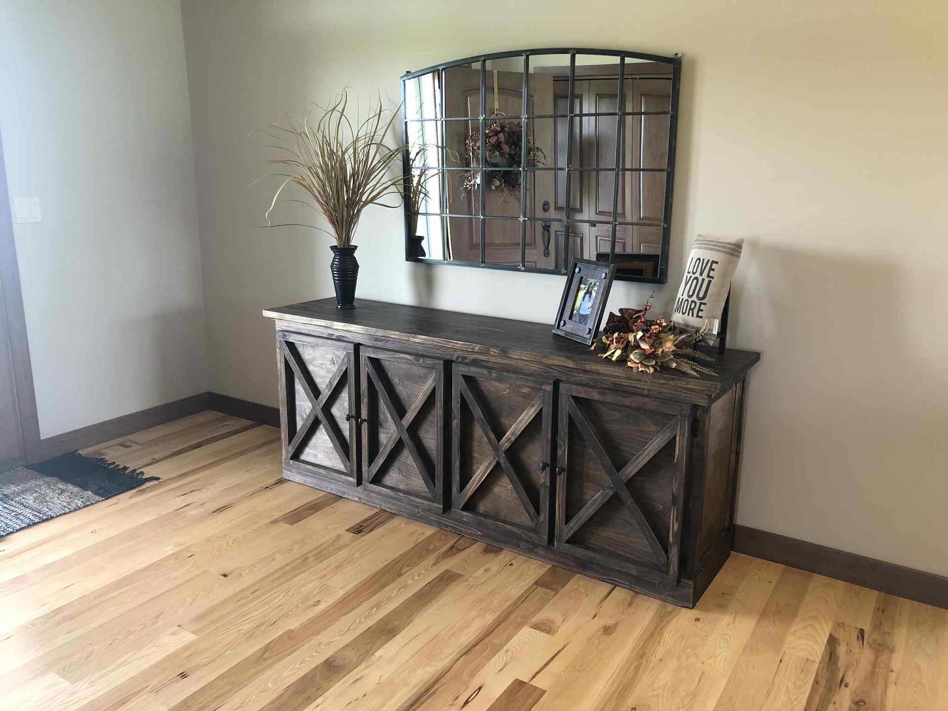Wooden sideboard with an X design, topped with decor and a mirror on a light wood floor.