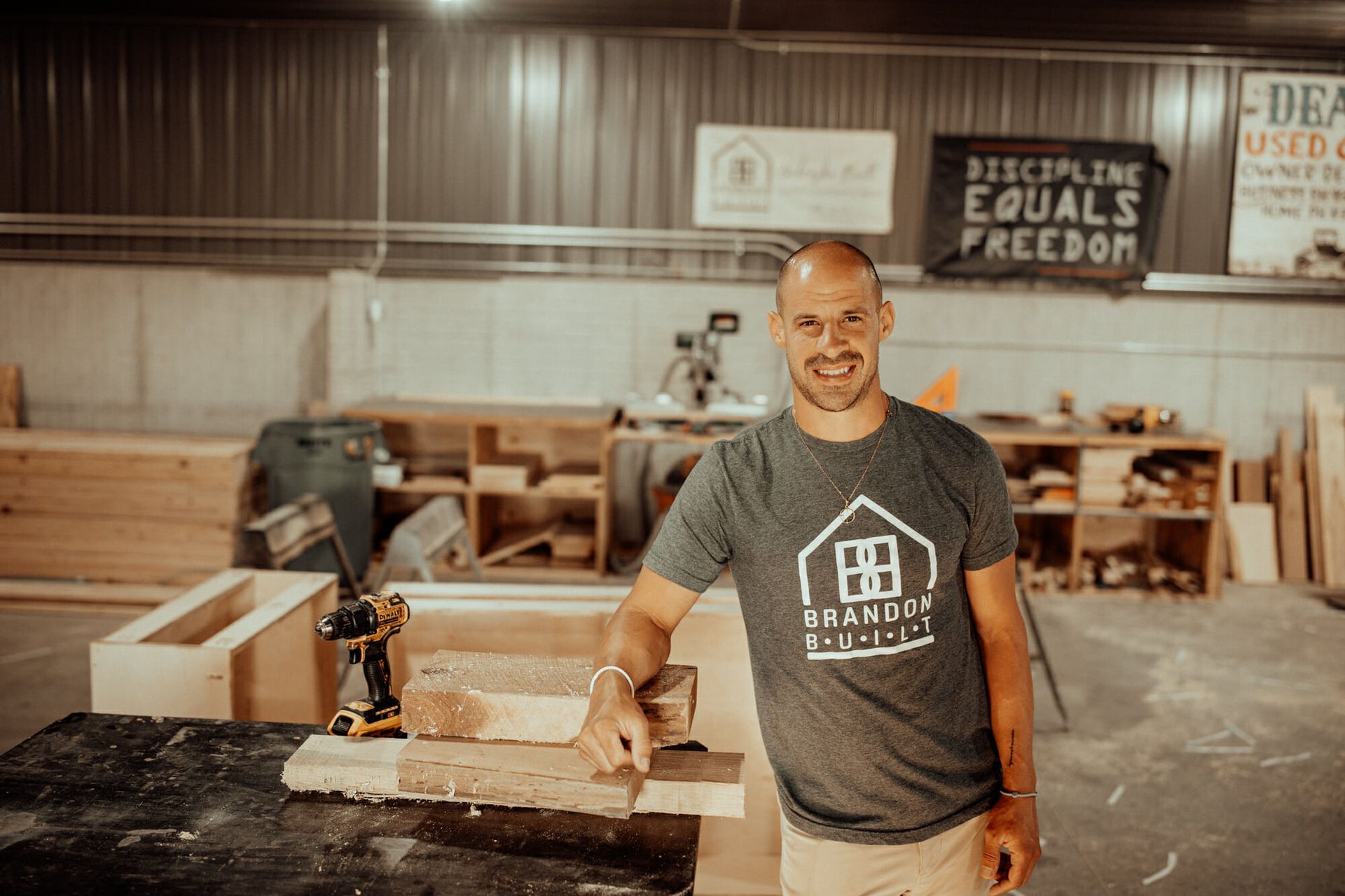 Man in workshop, smiling, leaning on wood. Sign reads