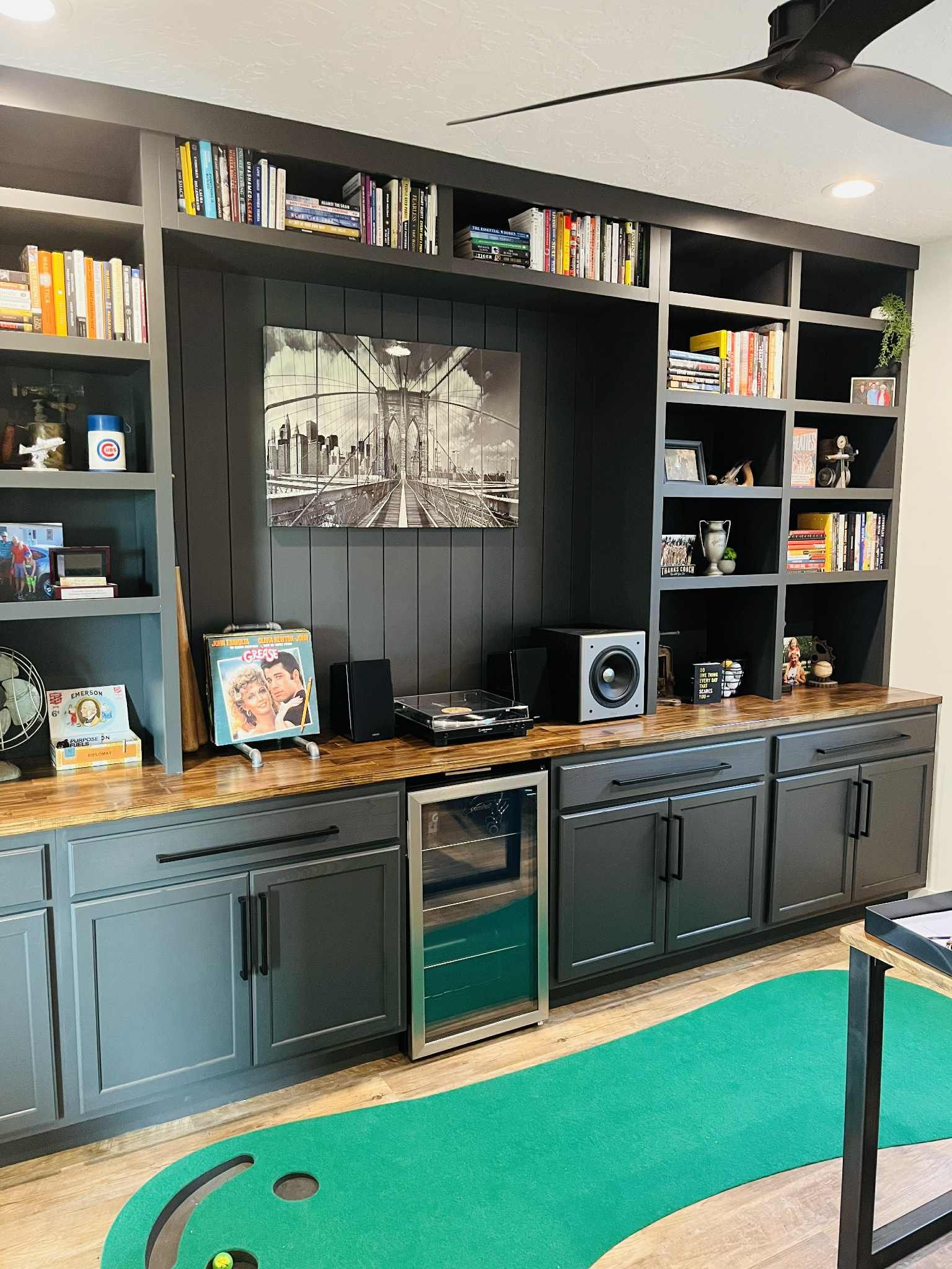 Built-in dark gray home office shelving with books, art, and a wine fridge. Includes a mini golf putting green.