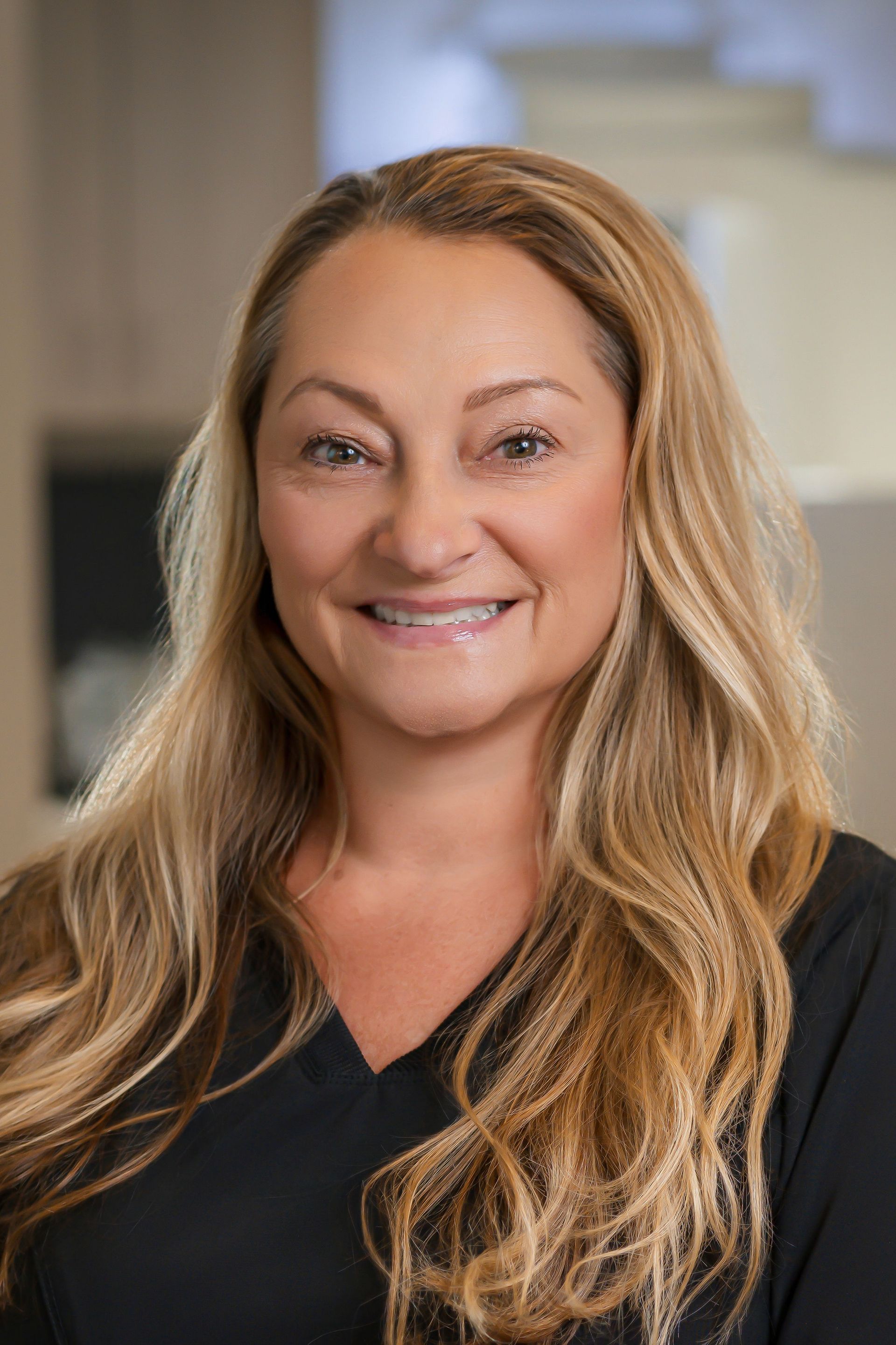 Woman with long blonde hair smiles, wearing a black top, in a well-lit office setting.