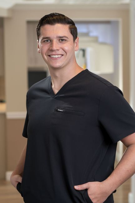 Man in black scrubs smiles, hands in pockets. Indoor setting, well-lit.