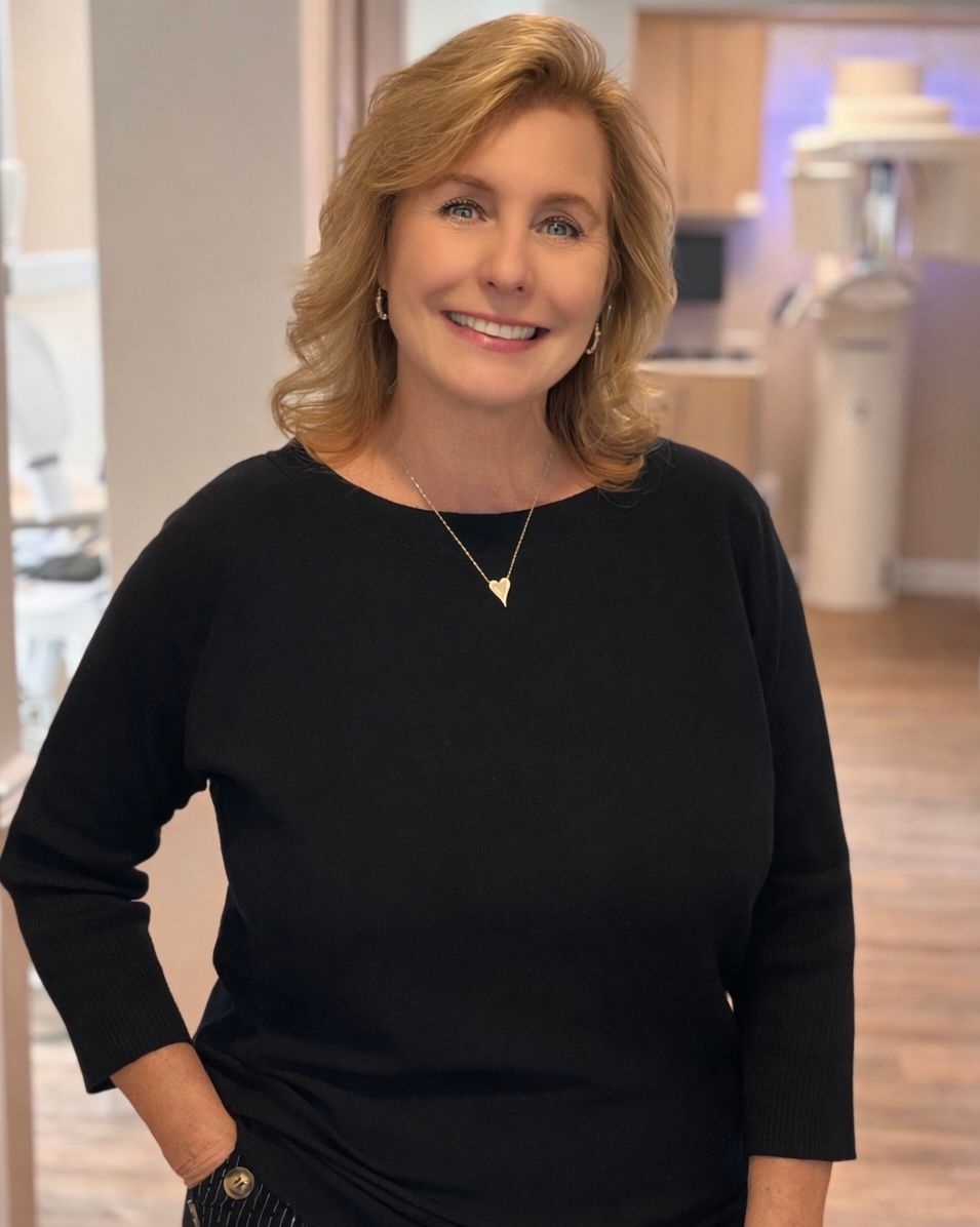 Woman in black top smiles, in dental office, hand in pocket.