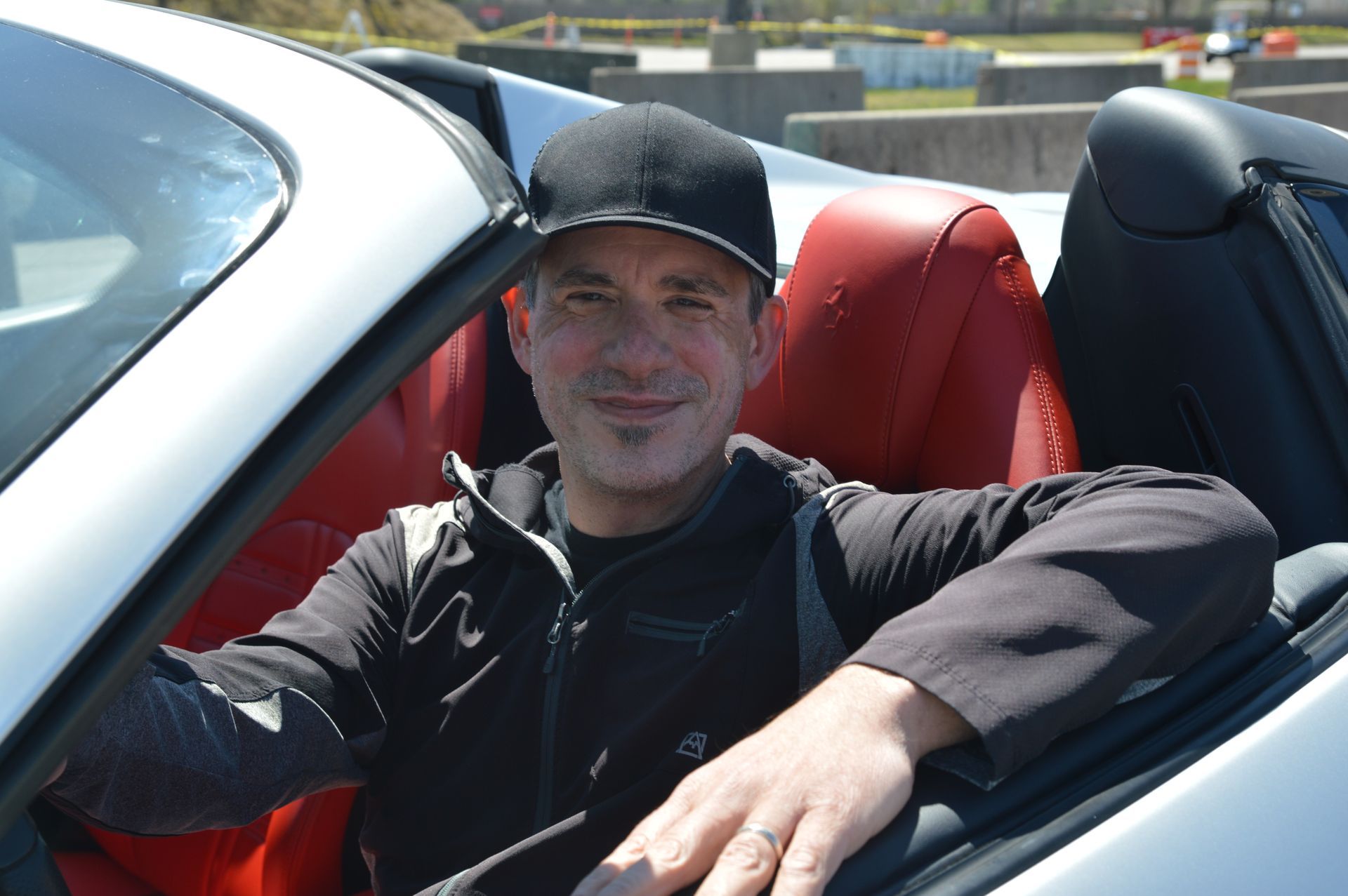 Man in black cap and jacket in a white convertible car with red seats, smiling.
