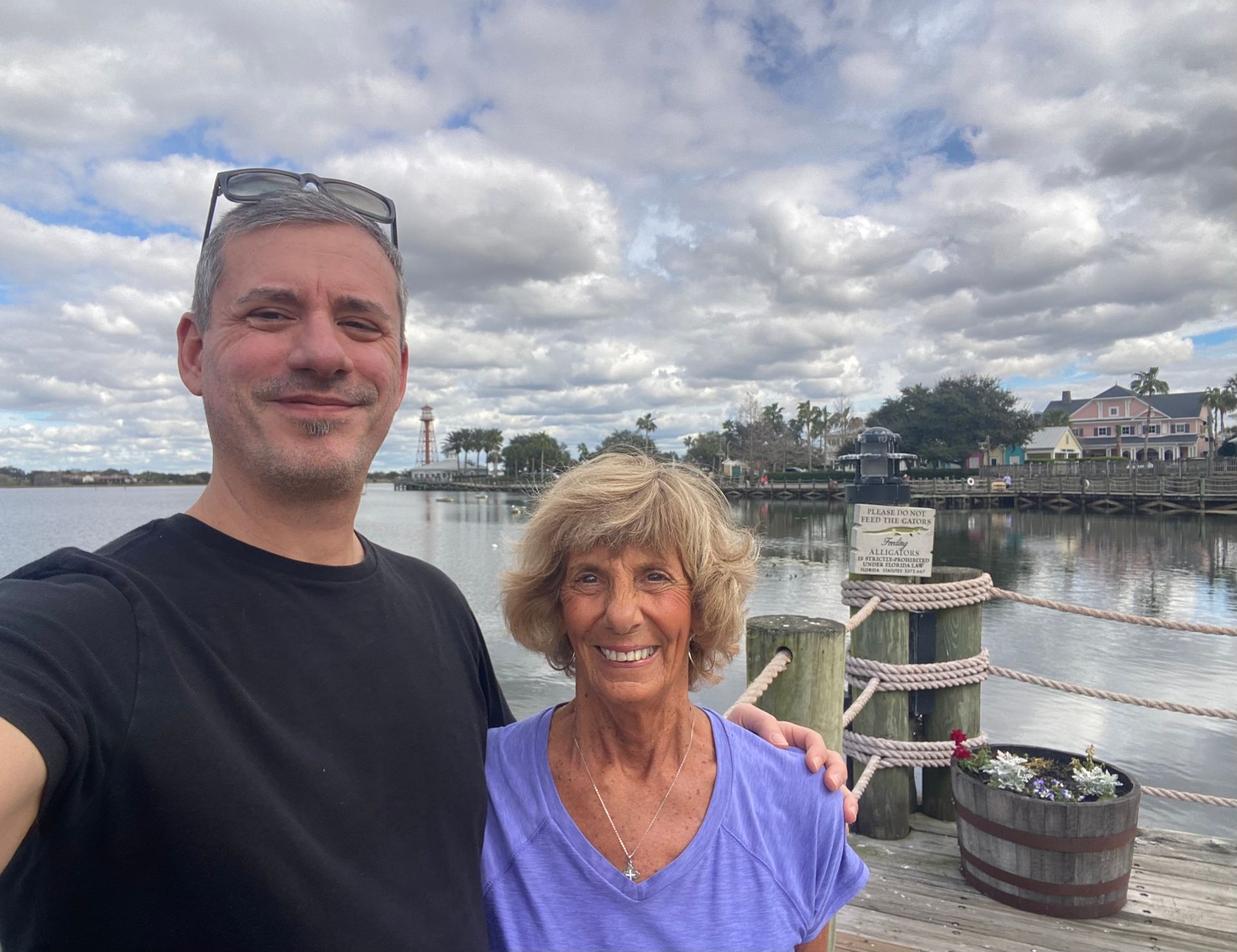 Man and woman on a dock taking a selfie, waterfront with lighthouse in background. Cloudy sky.