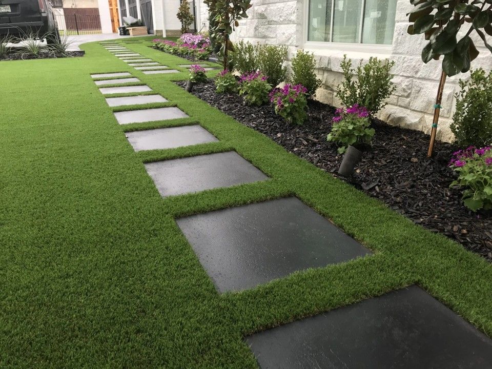 A path of square black stepping stones through green artificial grass in front of a house.