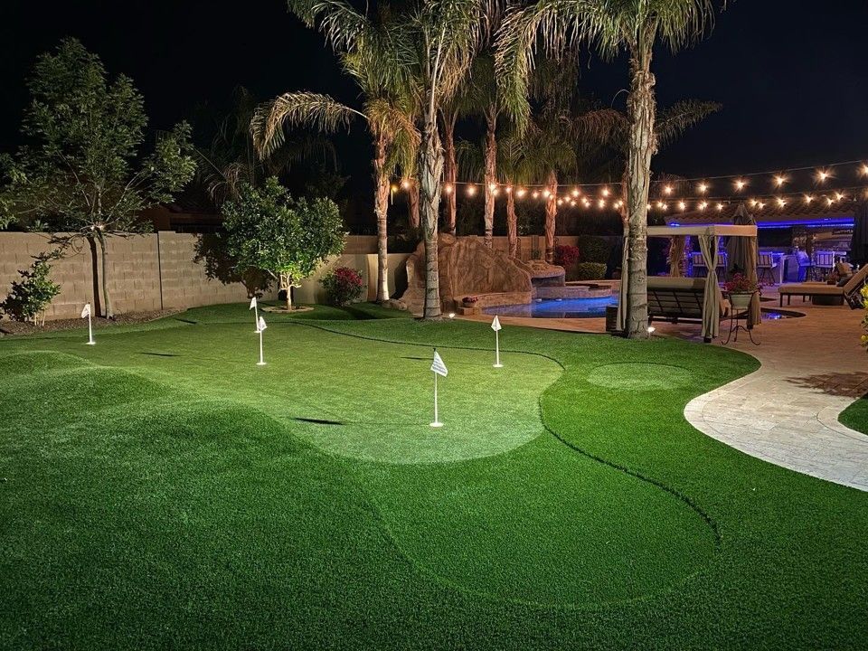 Night view of a backyard putting green with flags, string lights, and a pool.