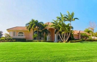Large pink house with tile roof, palm trees, and green lawn under blue sky.