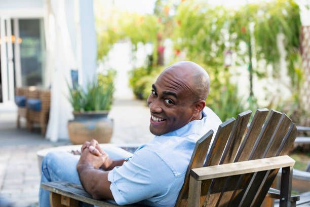 Bald man smiles while sitting in a wooden chair outdoors; light blue shirt and jeans.