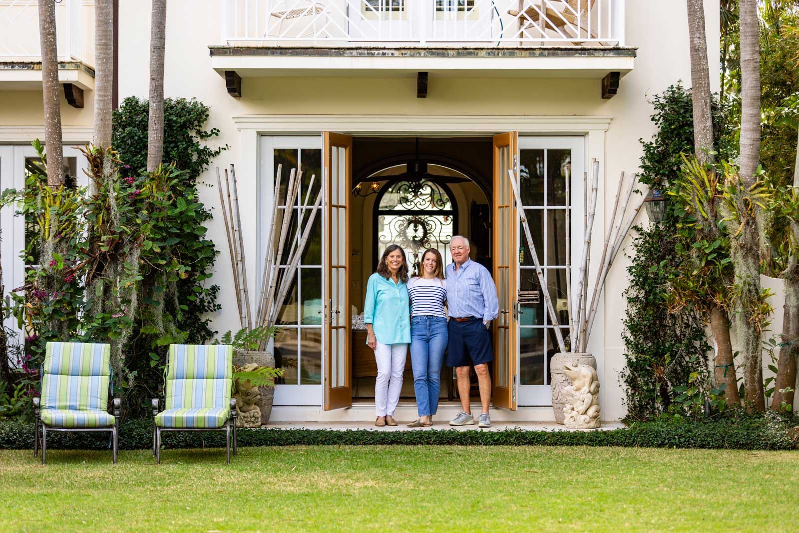 Three people stand in a doorway of a white house. Two chairs sit on the grass. Three people stand in a doorway of a white house. Two chairs sit on the grass.