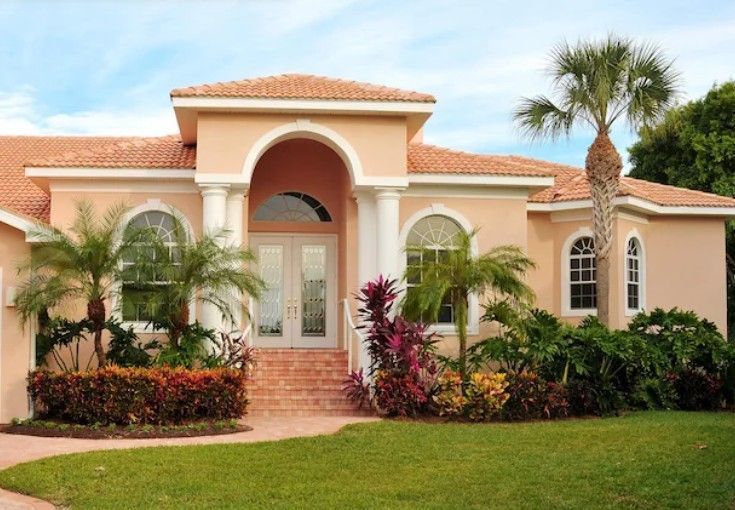 Beige stucco house with red tile roof, arched entryway, palm trees, and lush landscaping.