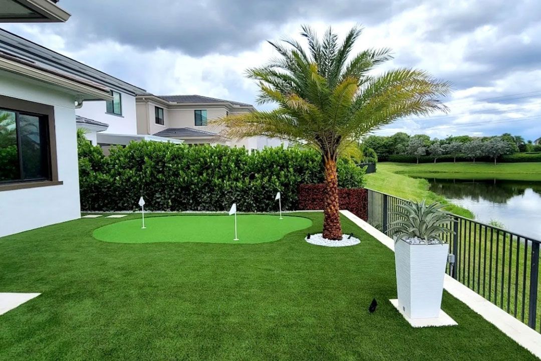 Backyard with putting green, palm tree, lake, and white and gray buildings.