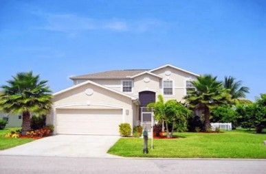 Two-story house with beige siding, a two-car garage, palm trees, and a green lawn under a blue sky.