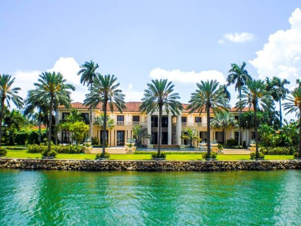 Large waterfront mansion with palm trees and green water.