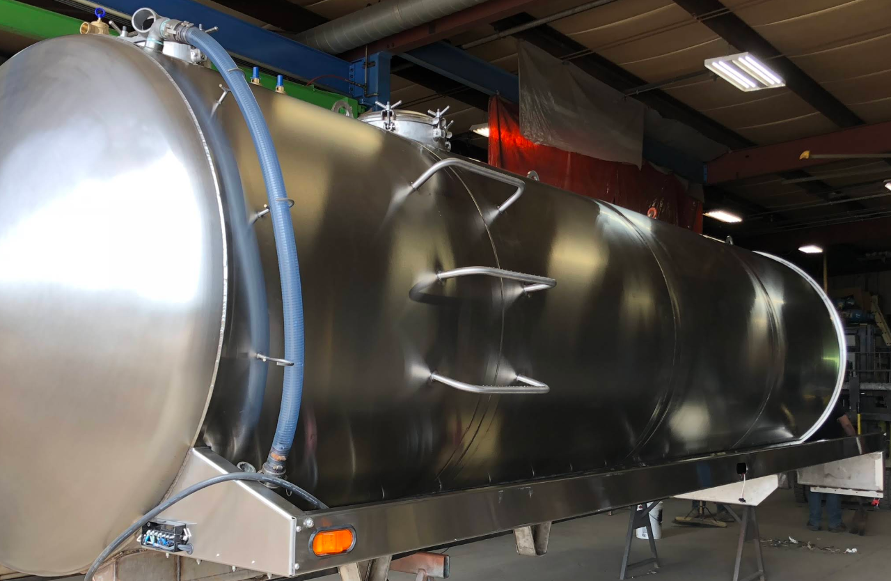 A large, shiny stainless steel tank, likely for transporting liquids, sits in a workshop. It has two round ends and is mounted on a frame.