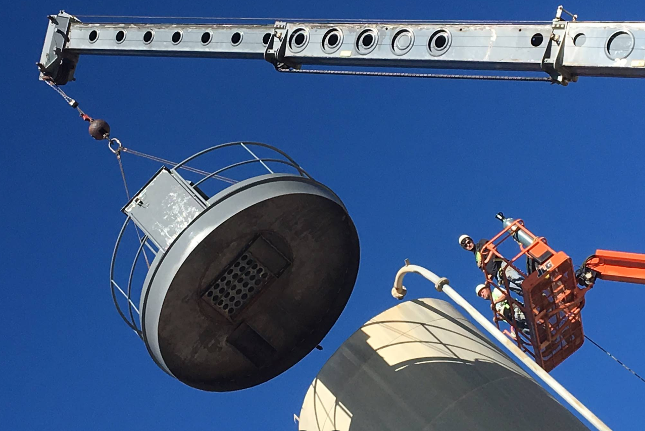 A crane lifting a large, circular metal structure above a cylindrical tank. A worker in an orange lift truck is nearby.