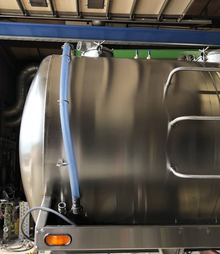 Shiny, silver tanker with a blue hose attached, possibly for transporting liquids. It is mounted on a trailer, likely in a factory setting.