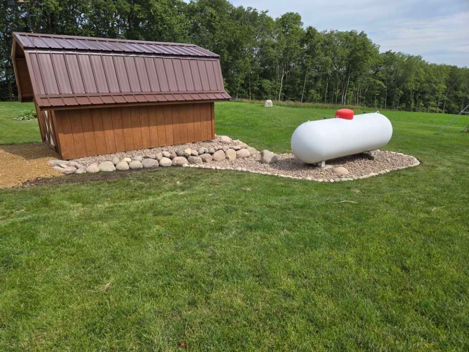 A propane tank is sitting next to a wooden shed in a grassy field.