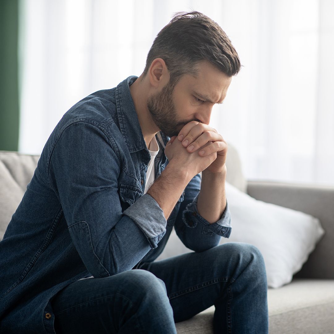 Mann i dongeri, i en sofa, hodet i hendene, ser ned, bekymret uttrykk.