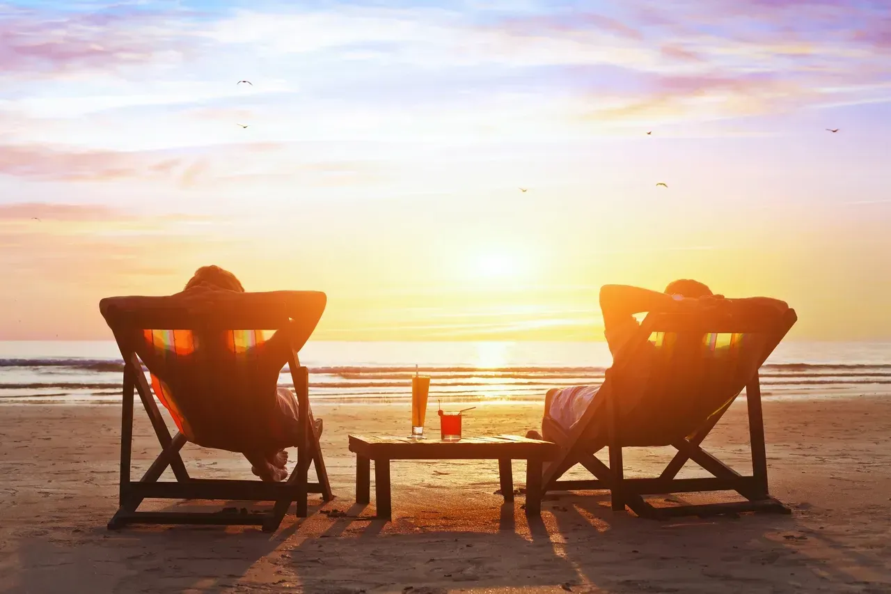 Two people are sitting in lawn chairs on the beach at sunset.
