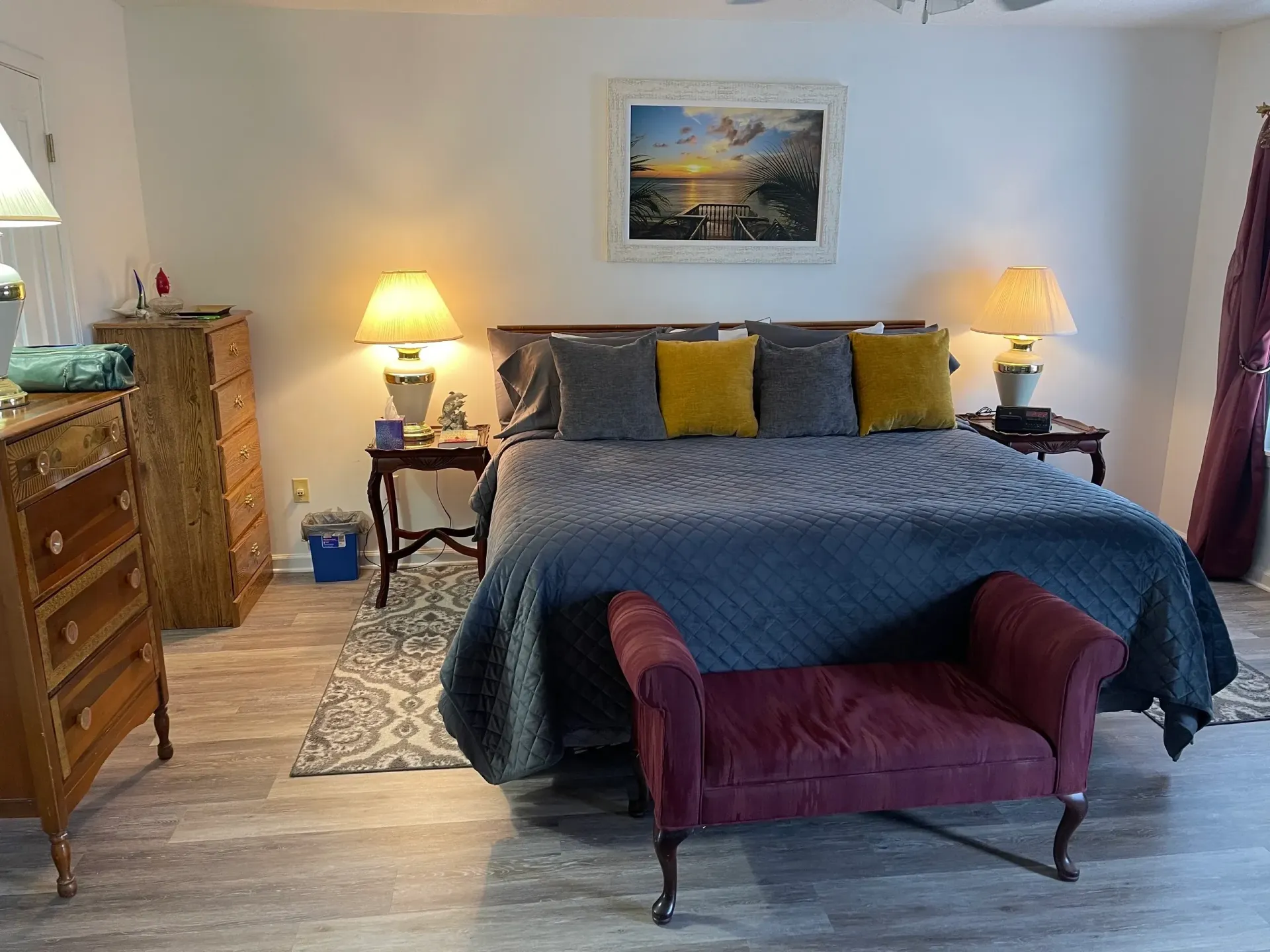 A bedroom with a bed , dresser , lamps , and a bench.