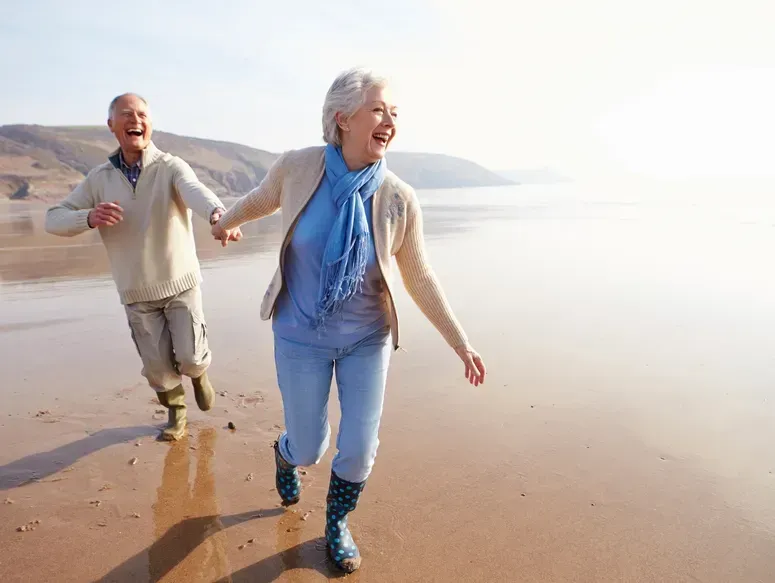 An elderly couple is running on the beach holding hands.