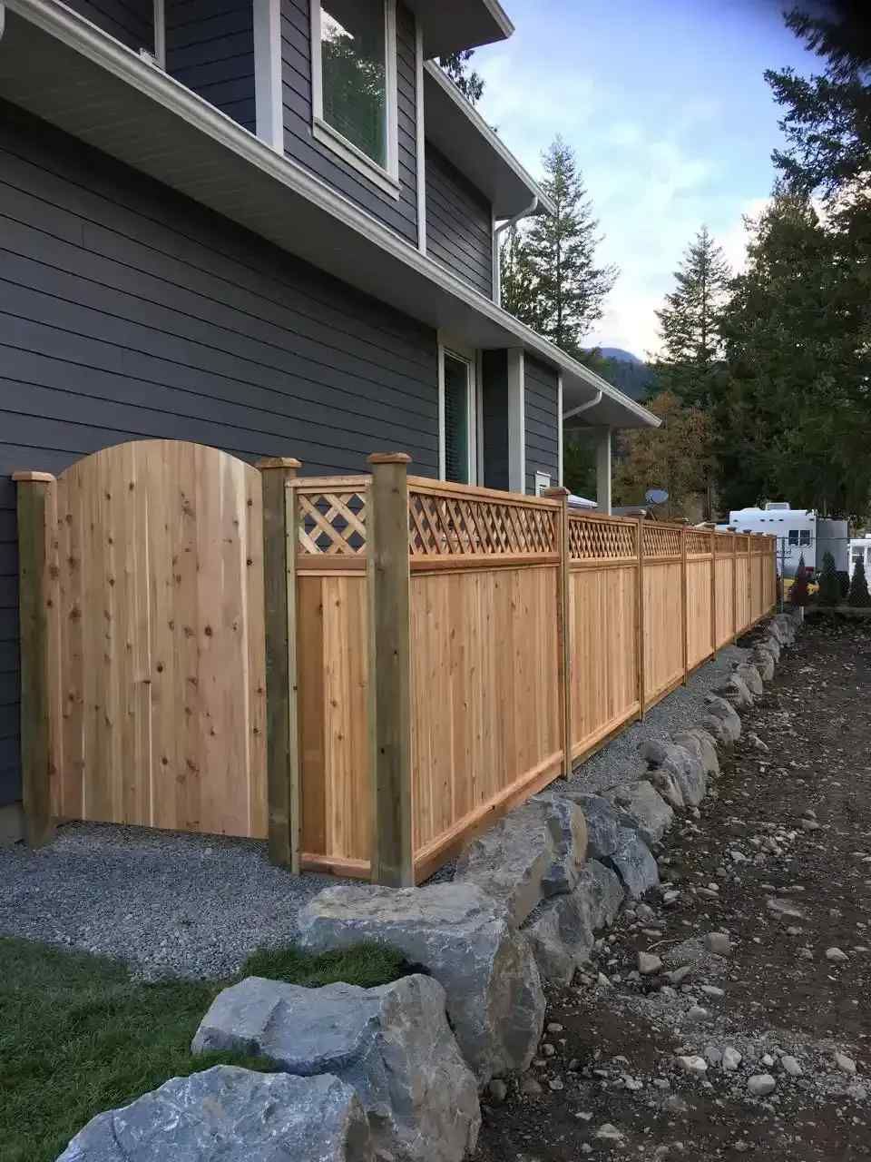 Wooden fence alongside a grey house with decorative lattice and arched gate, on a rocky base.