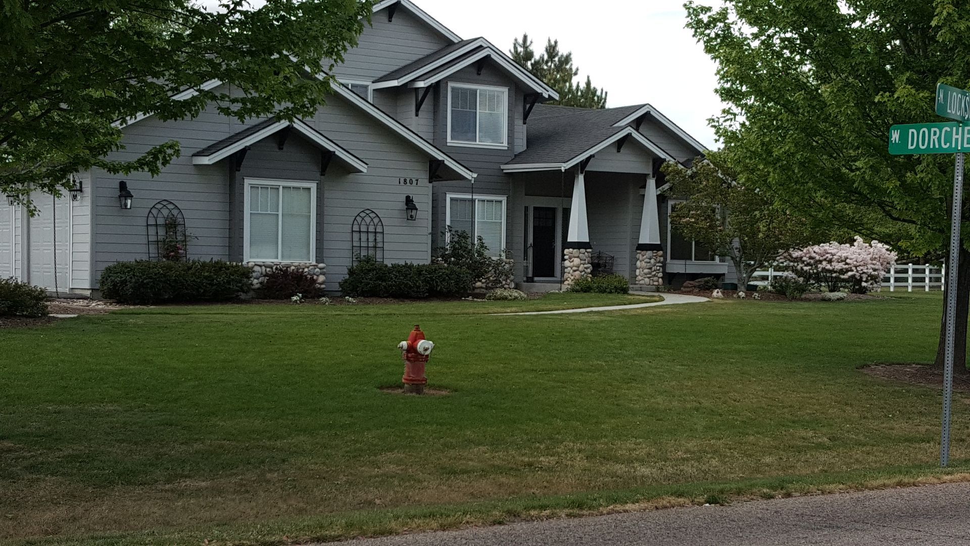 A gray two-story house with a green lawn and a red fire hydrant in front. A street sign reads 