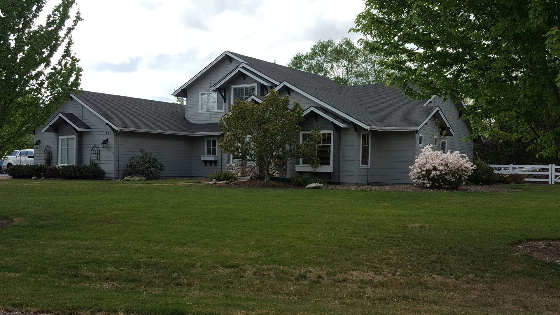 Gray two-story house with a dark roof, surrounded by green lawn and trees, under a cloudy sky.