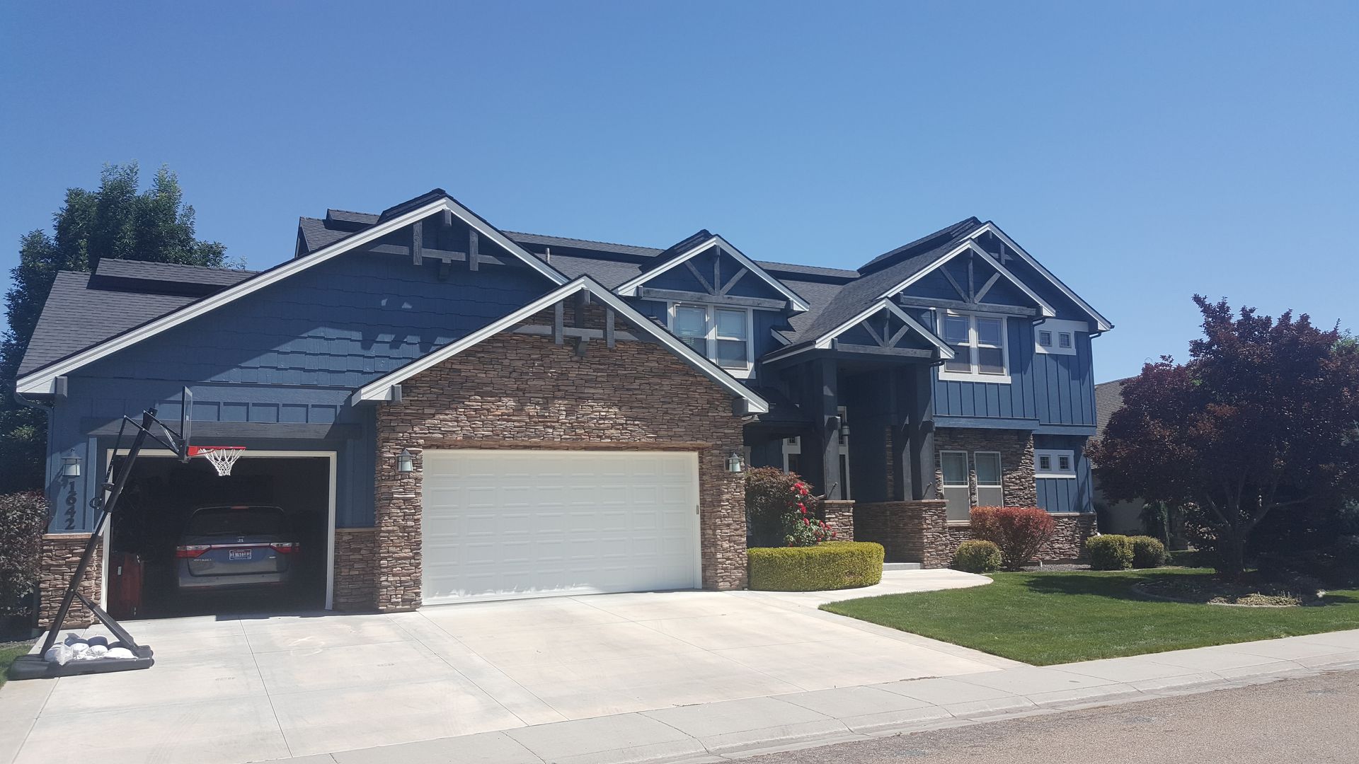 Blue and stone-clad two-story house with a two-car garage, basketball hoop, and well-manicured lawn on a sunny day.