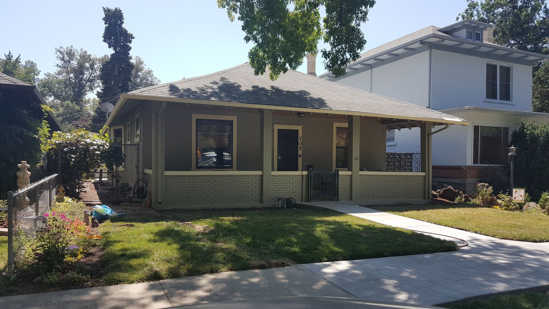 Small, tan house with a porch. Green grass, sidewalk, and neighboring white house.