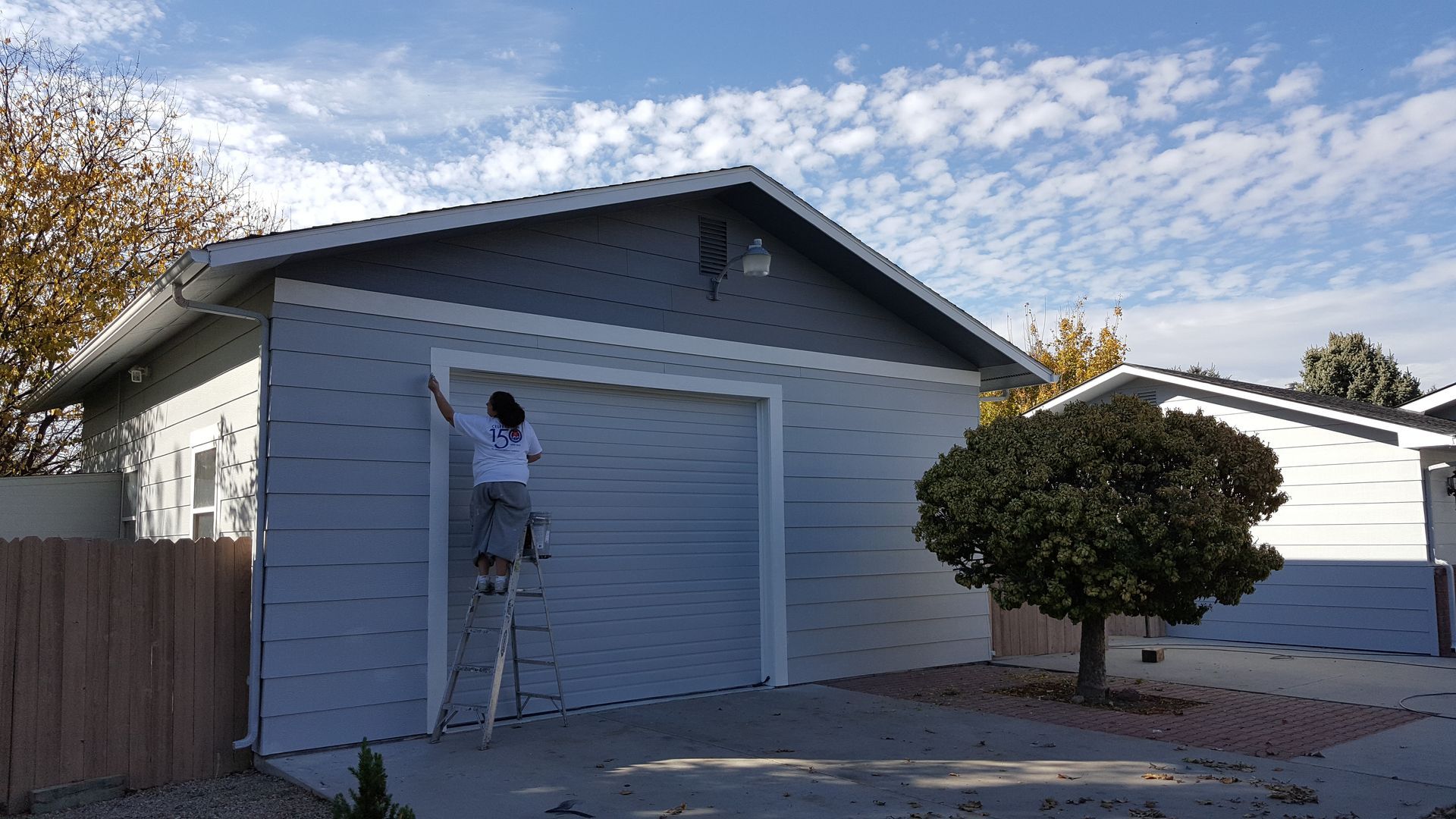 Person on ladder painting a gray garage door with white trim under a partly cloudy blue sky.