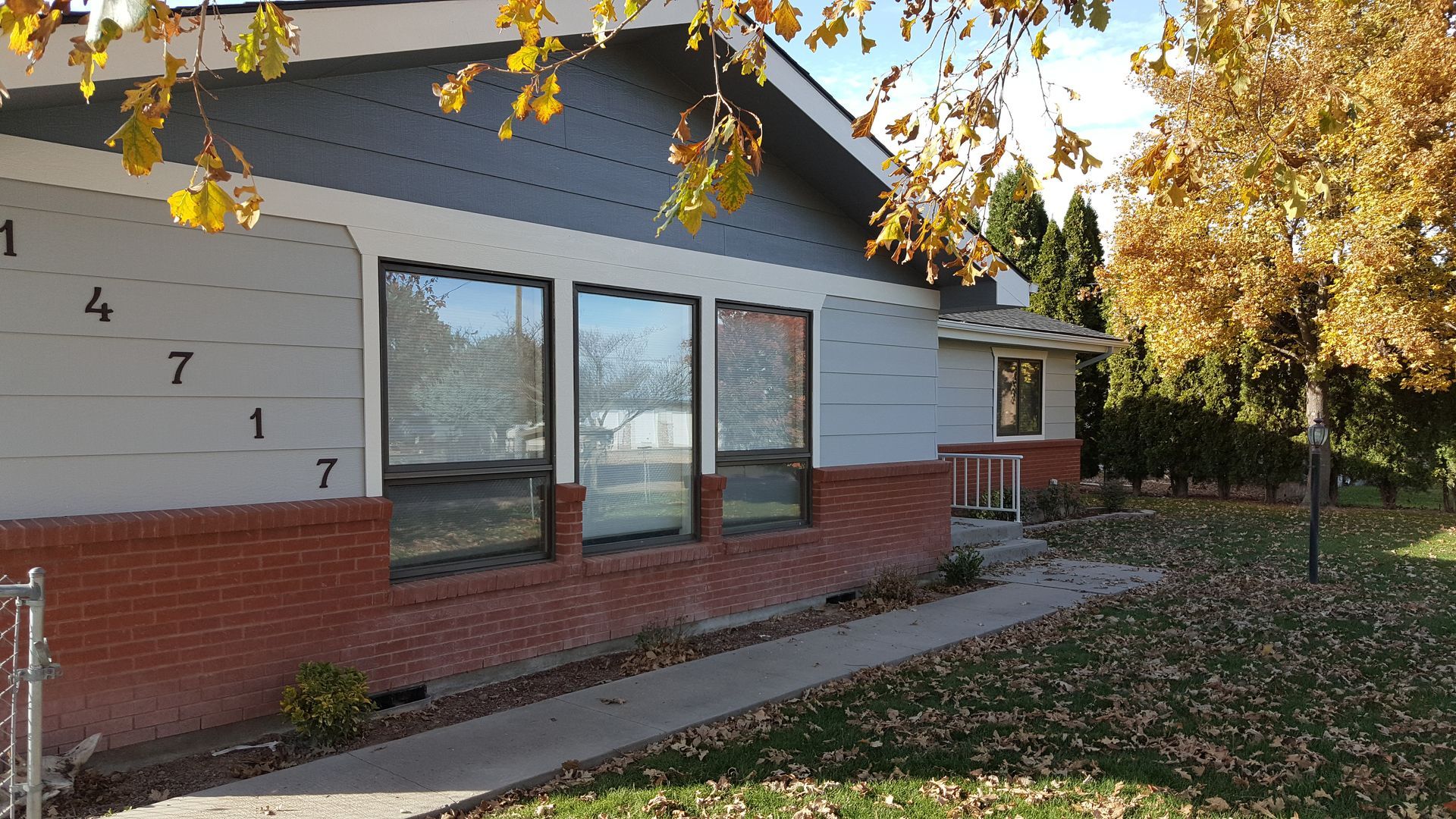 A light gray house with a brick base and large windows. The house number 4717 is visible. There's a walkway and trees with fall foliage.