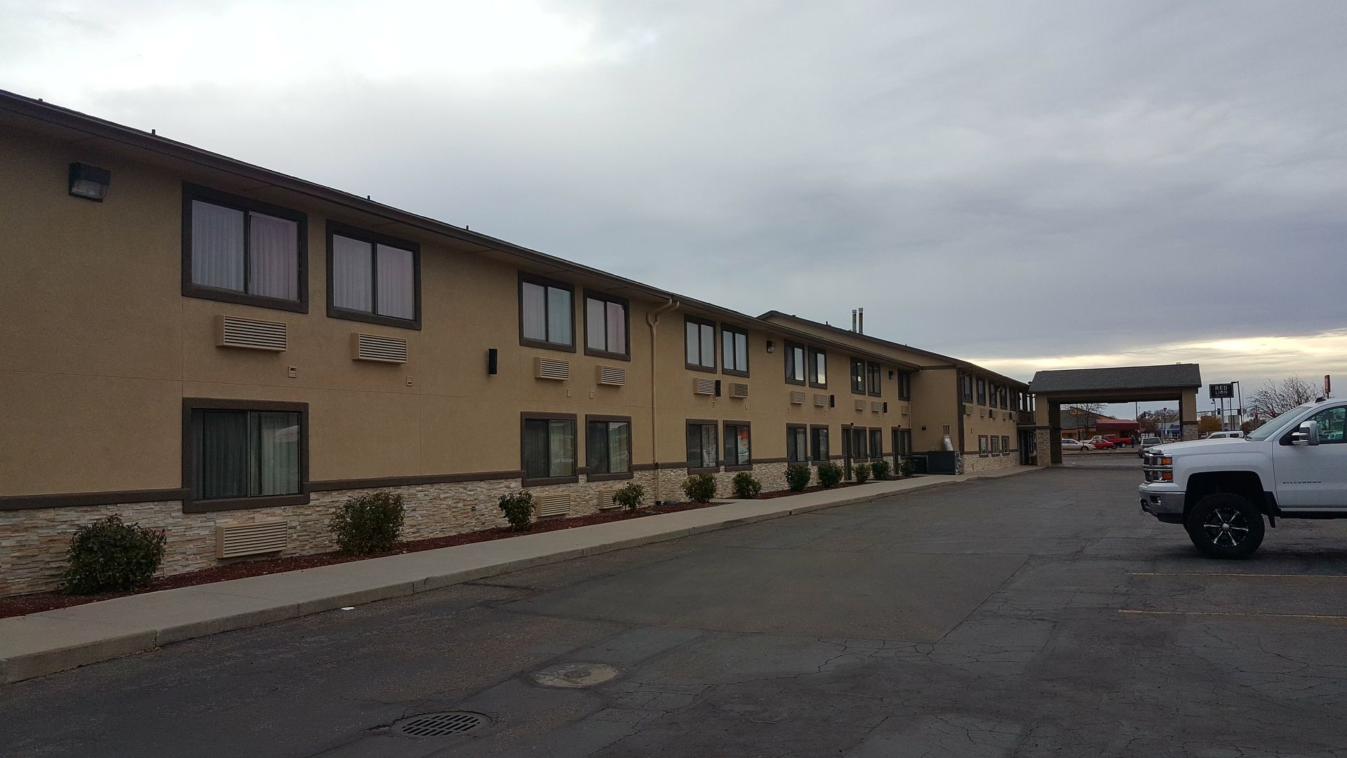 A beige motel with two stories, brown window frames, and a paved parking lot under a cloudy sky. A white truck is parked on the right.