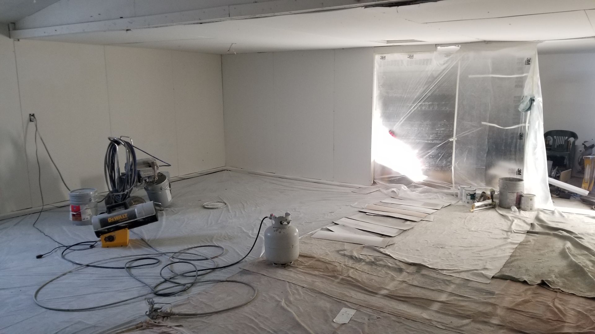 An interior room being painted, covered in protective sheeting and drop cloths. Paint equipment and supplies are visible.