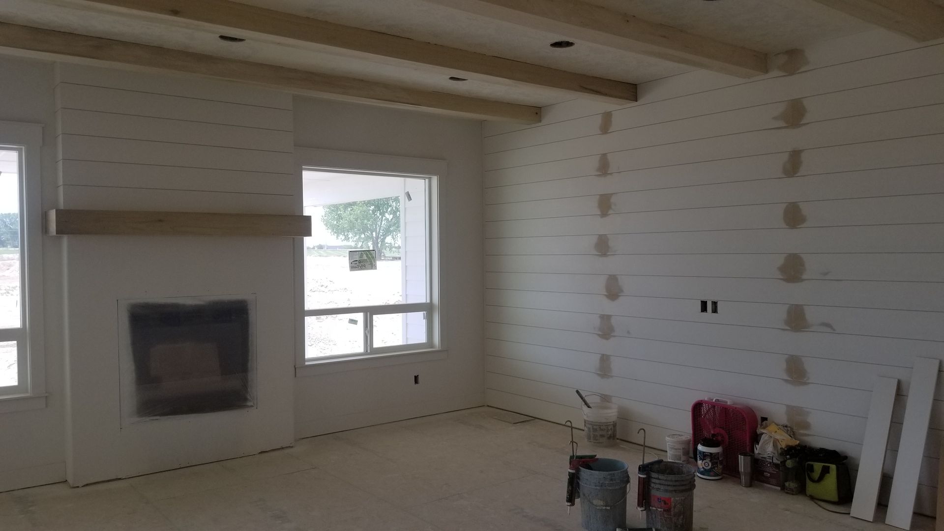Interior room with white plank walls, a fireplace, windows, and wooden beams across the ceiling. Paint supplies are in the corner.