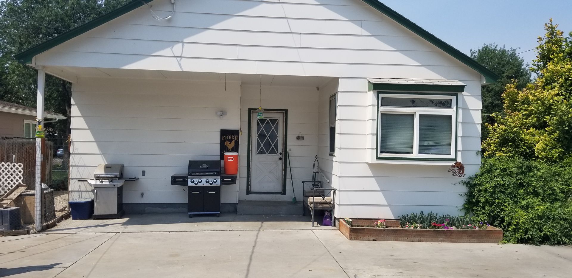 White house with a porch, grill, and garden bed. Door with glass panels and a window with a sunshade.