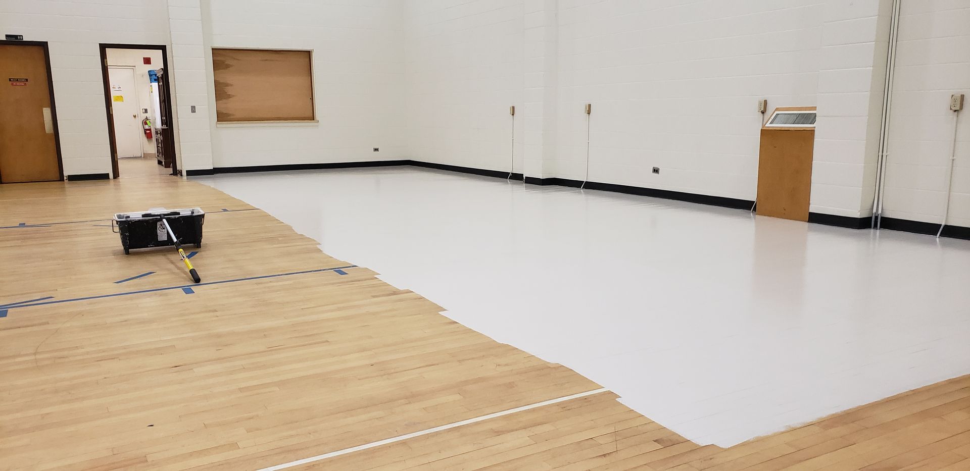 A gym floor being repainted; one side is wood, the other is white. A machine in the middle of the floor is painting the white section.