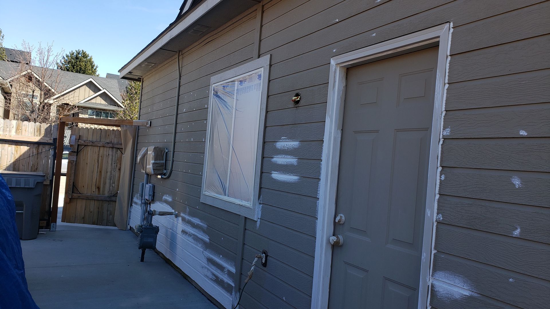 Side of a house with light gray siding, a door, and a window covered in plastic. Exterior shows signs of repair work.