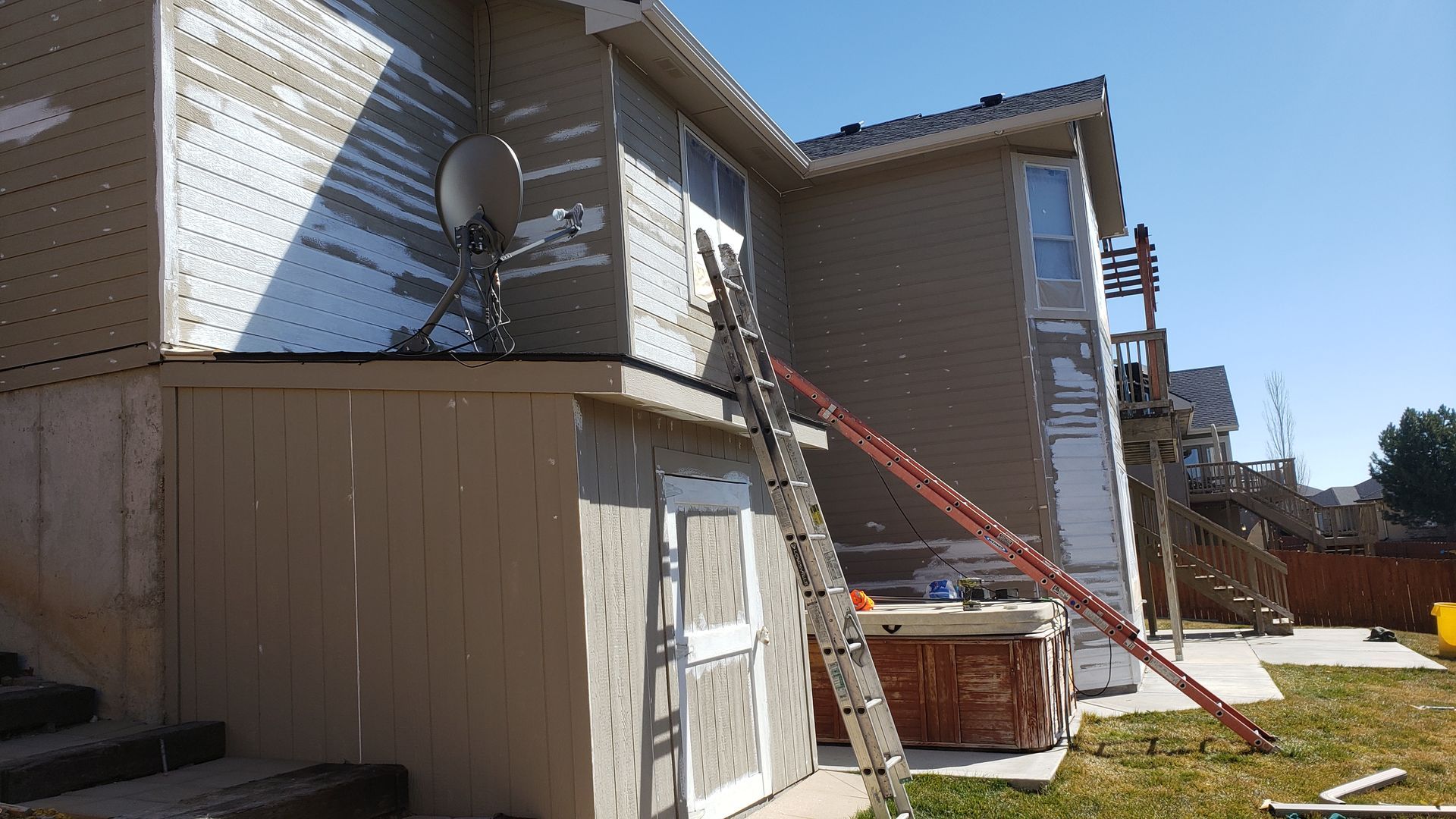 Side of a two-story house undergoing siding repair, with ladders, satellite dish, and hot tub visible.