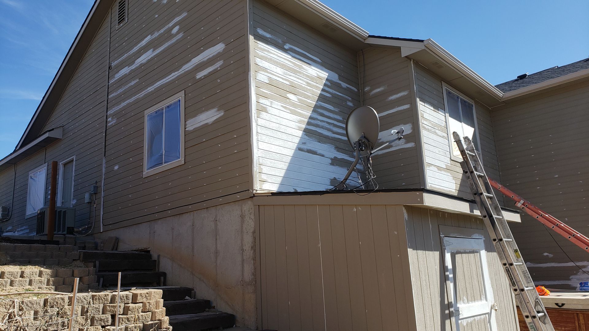 A tan house with visible repairs on the siding and a ladder set up next to a door.