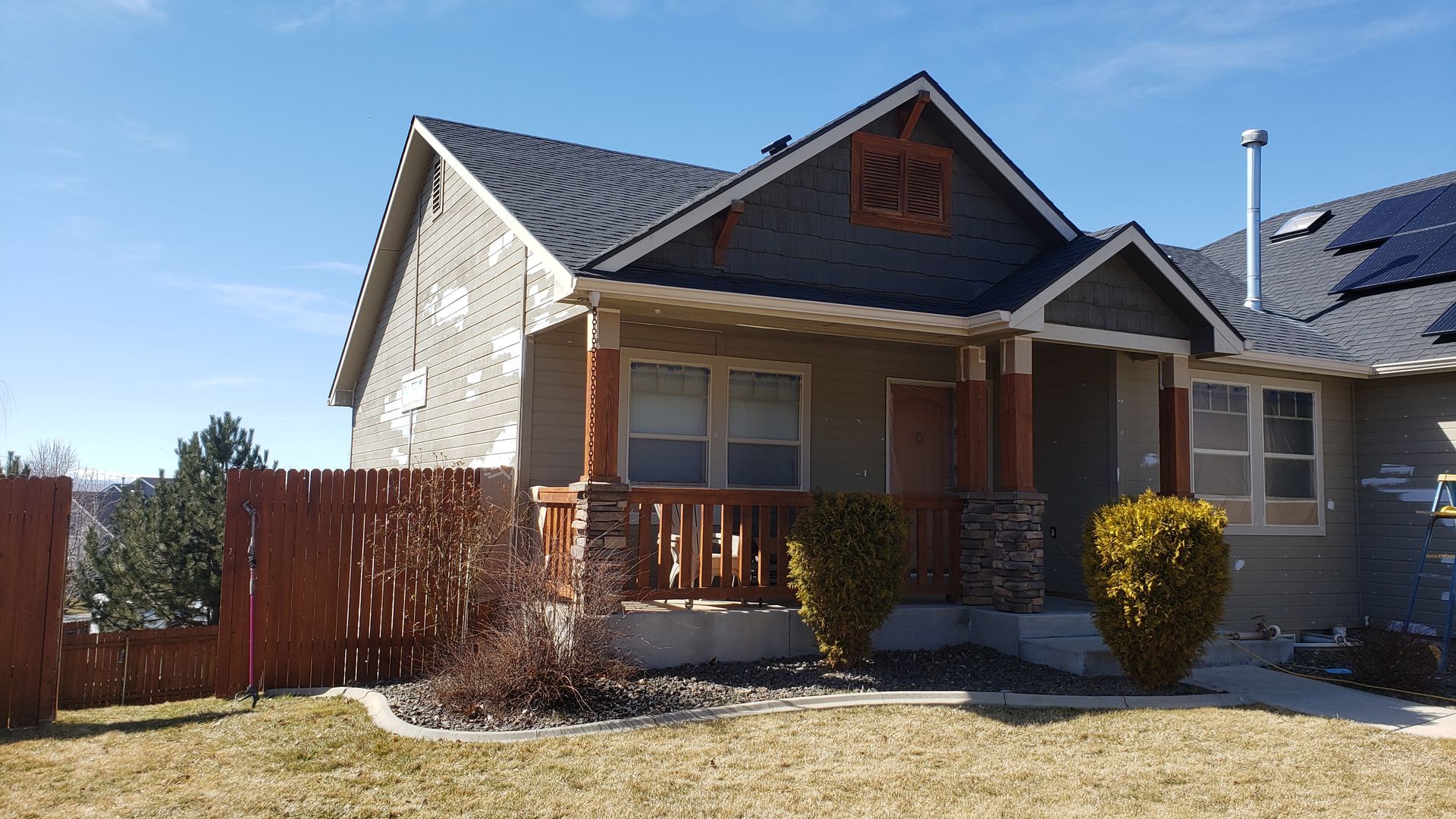 A tan house with a porch, dark roof, and brown fence. A sunny day with solar panels on the roof.