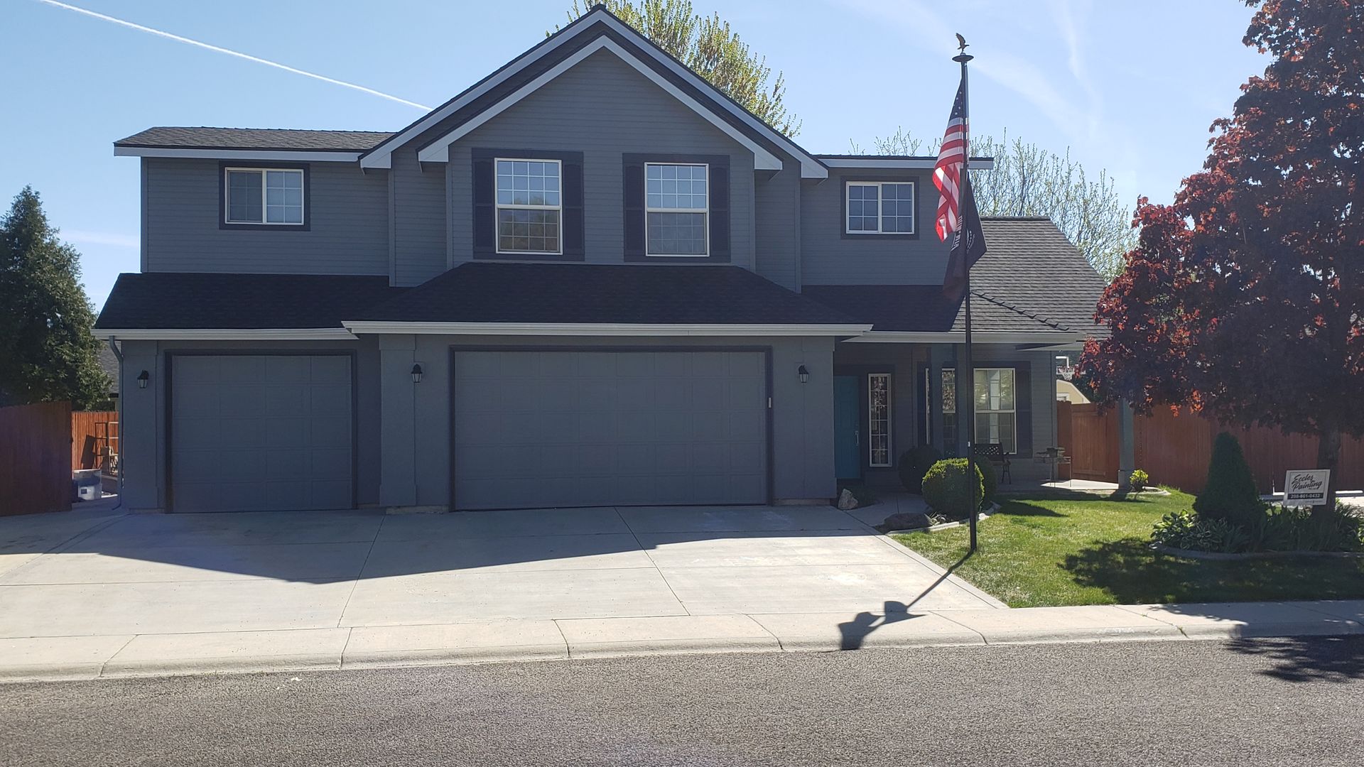 Two-story gray house with black trim, two garage doors, and an American flag. The house has a front yard with a driveway.