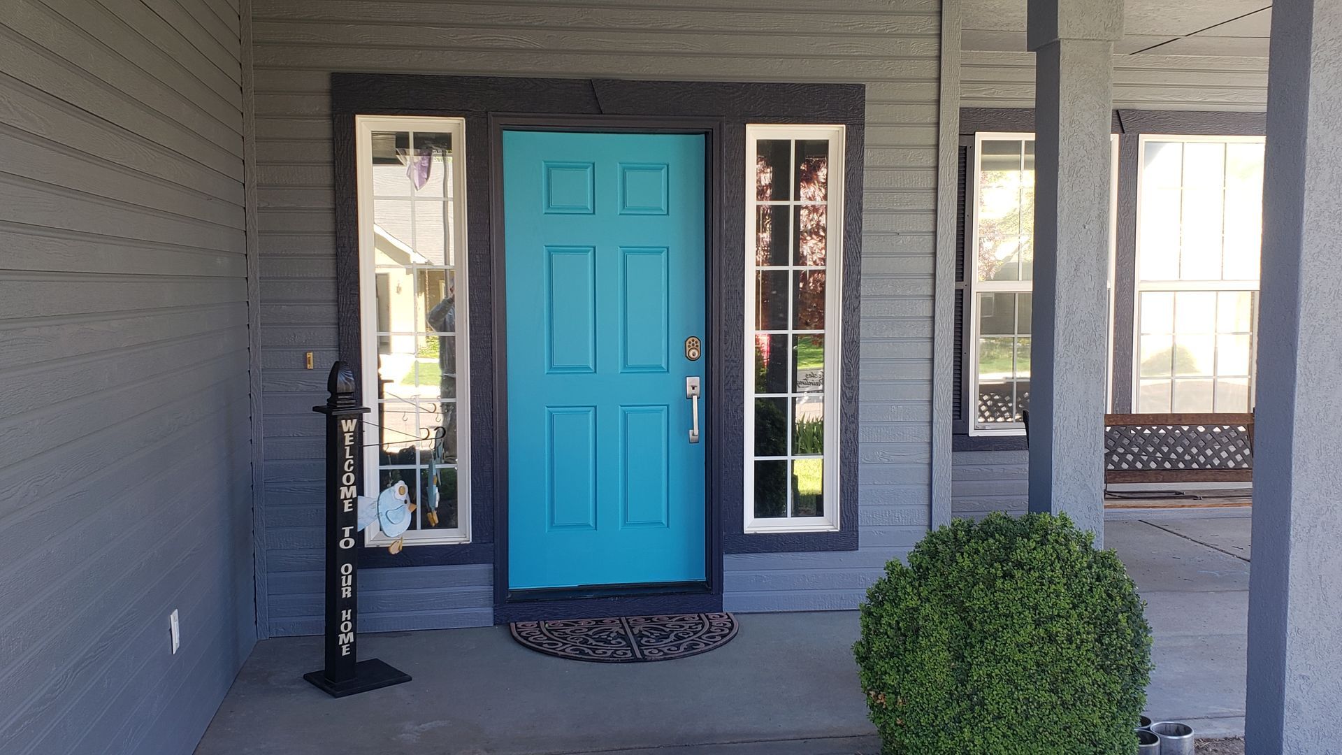 Teal front door with white-trimmed sidelights on a gray house. A round bush sits to the right of the door.