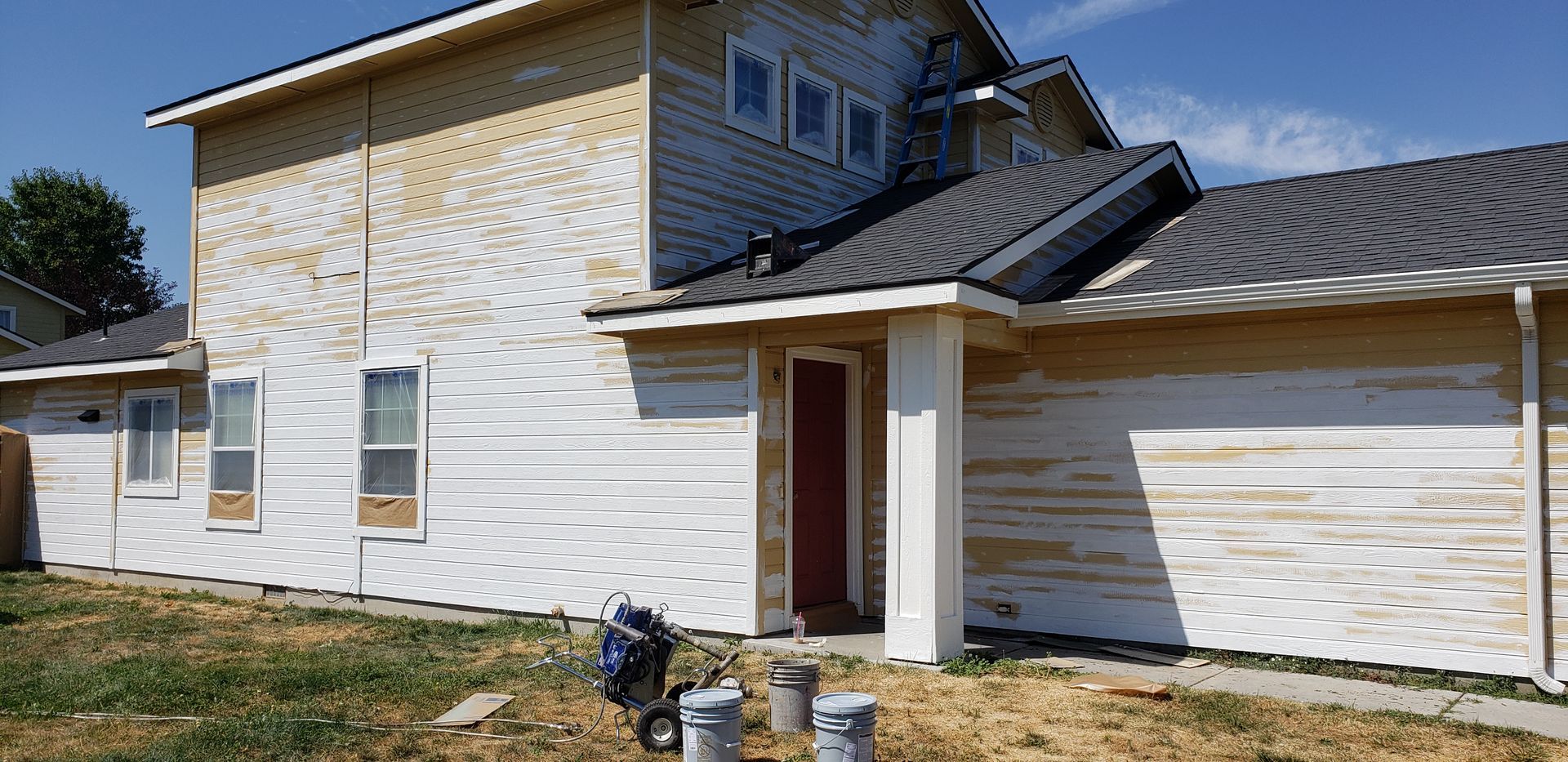 A house under construction with exposed siding. It has a red door, a dark roof, and sits on a grassy area under a blue sky.