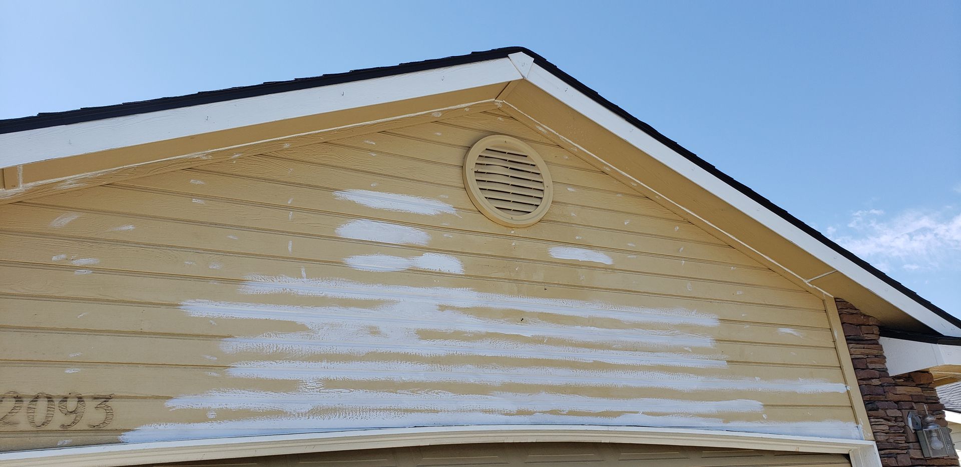 Close-up of a house exterior with yellowish siding, a black roof, and white splotches, likely from paint. A blue sky is in the background.