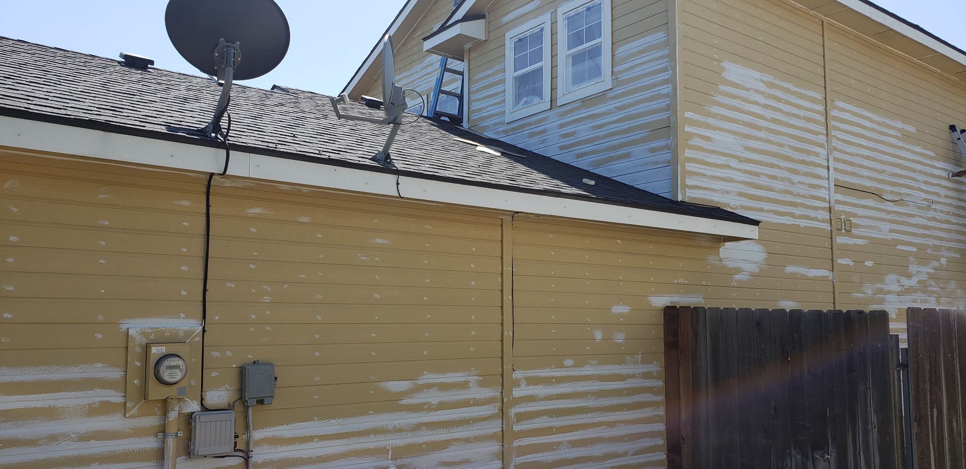 Exterior of a yellow house with visible paint peeling. A satellite dish is mounted on the roof, and a wooden fence is visible.