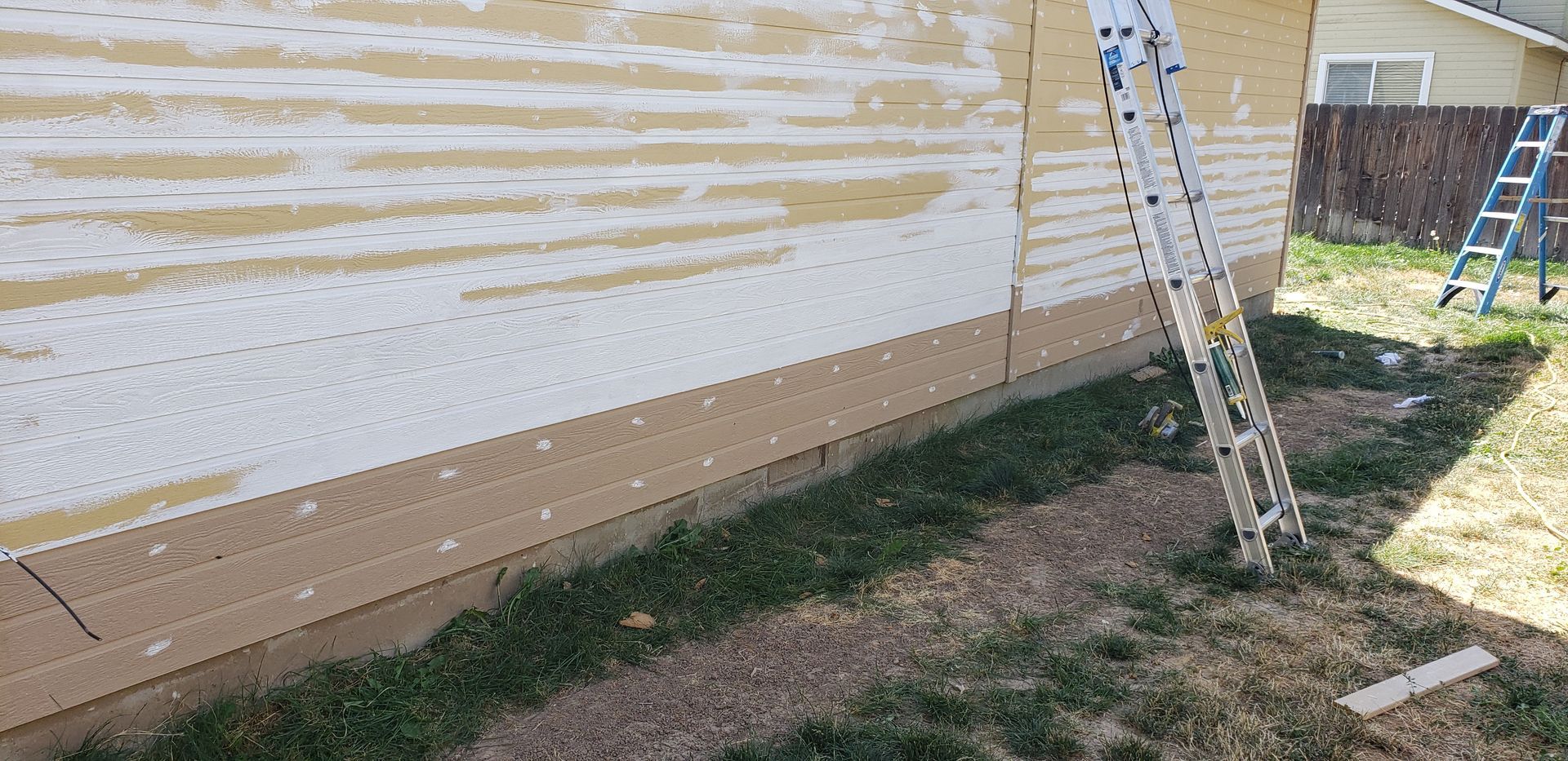 A house with new siding, a ladder, and a second ladder in the background. Green grass and gravel are in the foreground.